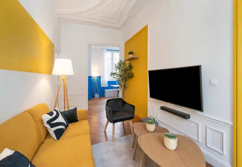 Bright, stylish living room with a mustard sofa, accent chair, wall-mounted TV and wooden nesting tables — modern decor with warm textiles and plants creating a welcoming atmosphere.