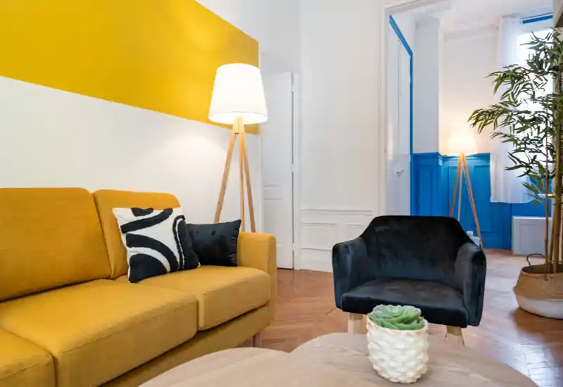Bright, stylish living room with a mustard sofa, dark accent chair, floor lamps and decorative plants, offering a cozy, modern sitting area.