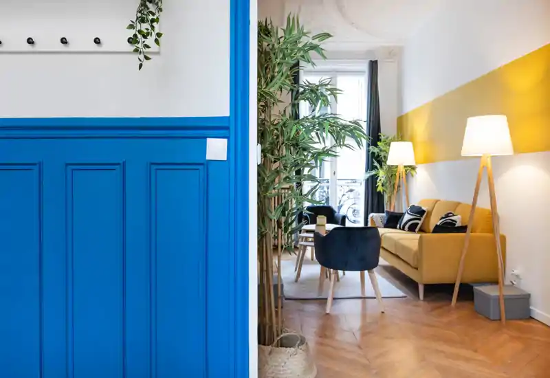 Stylish, well-lit living room with a mustard sofa, accent chairs and floor lamps; decorative plants and a balcony window create a bright, inviting space.