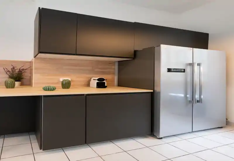 Modern compact kitchen corner with dark matte cabinets, a wooden countertop and backsplash, small coffee machine and a stainless steel side-by-side refrigerator — bright, tidy and stylish.