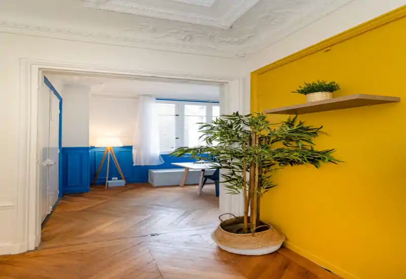 A bright, stylish living area with bold blue wood panelling and warm herringbone parquet. Natural light from a tall window, a floor lamp, and tasteful plants create a welcoming, well-styled space.