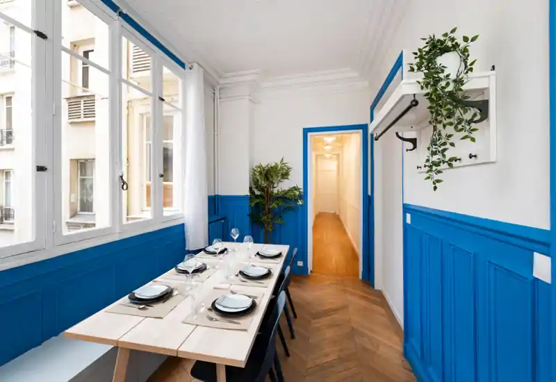 Bright, styled dining room with a long wooden table set for six, large windows providing natural light, and bold blue wall panelling for a modern, welcoming atmosphere.
