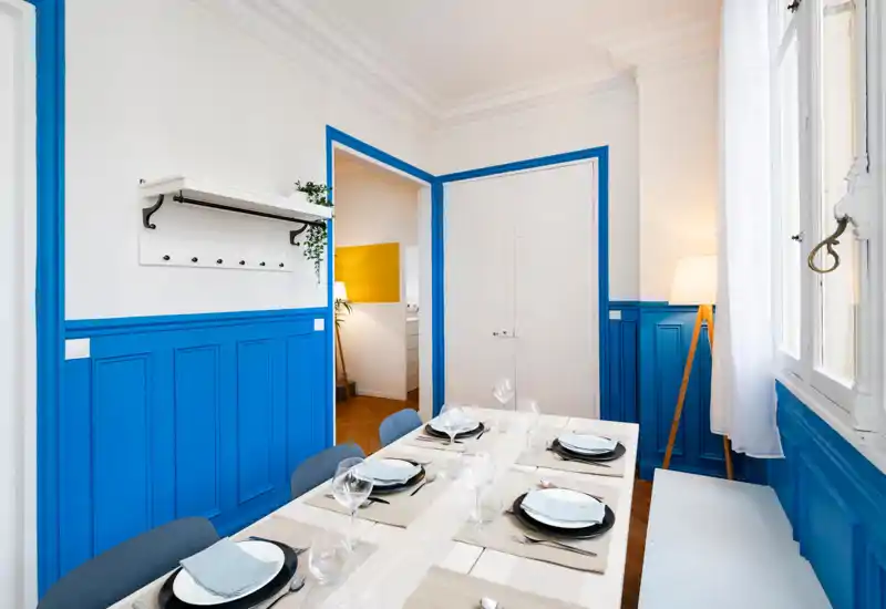 Bright, styled dining room with a set table for six, blue wainscoting and natural light from a large window creating a fresh, modern atmosphere.