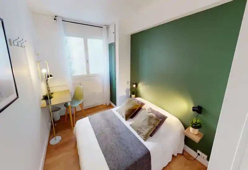 Sunlit, cozy bedroom with a green accent wall, comfortable double bed with neutral linens, bedside lights and a small desk by the window — ideal for relaxing or working.