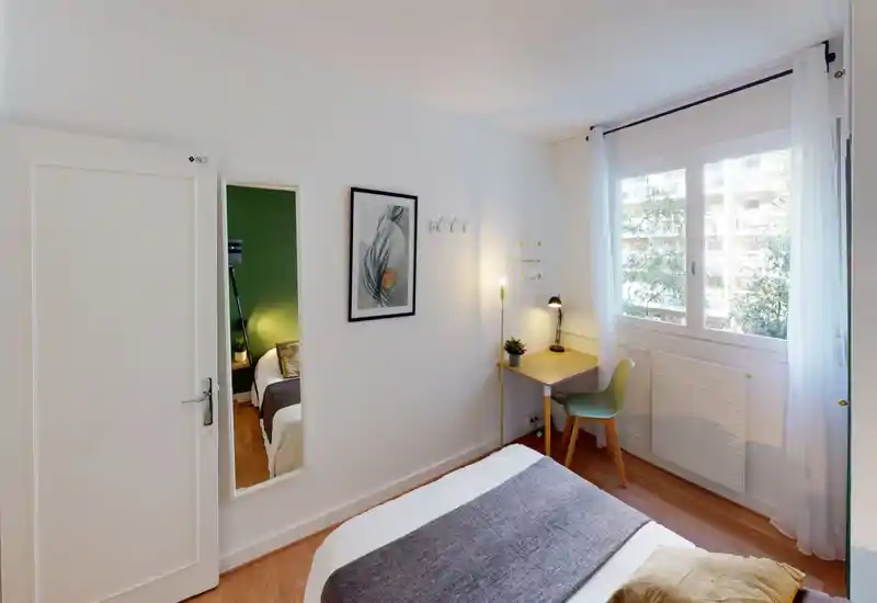 Bright, neatly styled bedroom with a visible double bed in the foreground, a small desk by the window and soft natural light that creates a welcoming atmosphere.