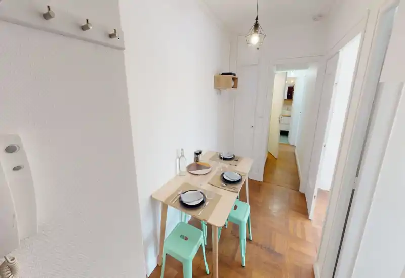 Compact dining area with a light wooden table set for four and pastel stools, bright white walls and warm parquet flooring — ideal for small apartments or breakfast nooks.