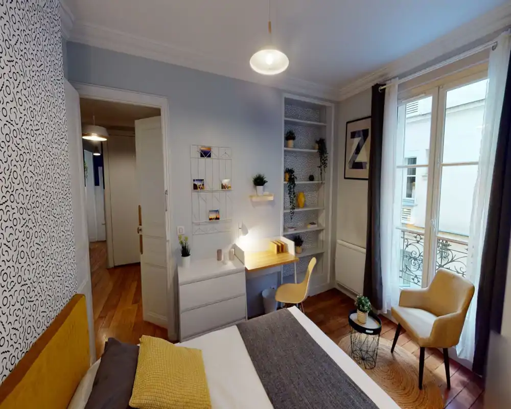 Camera da letto luminosa e arredata con letto ben visibile, una piccola scrivania, scaffali e una poltrona vicino alla finestra che crea un angolo lettura accogliente.