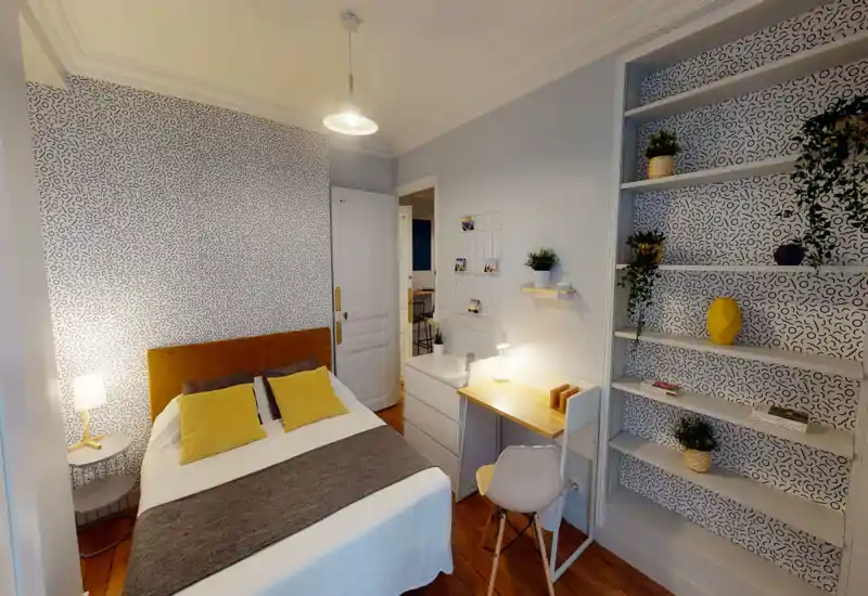 Camera da letto luminosa e alla moda con un letto matrimoniale, testiera calda e cuscini gialli coordinati. Mensole integrate, una scrivania compatta e carta da parati decorativa creano uno spazio accogliente e ben organizzato, ideale per una persona o una coppia.