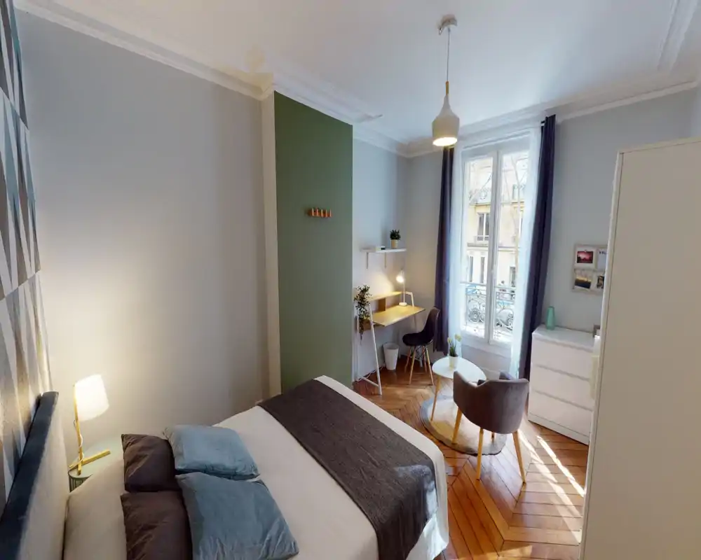 Camera da letto luminosa e di design con un ampio letto, pavimento in parquet, una postazione di lavoro vicino alla finestra e un angolo salotto accogliente — ideale per soggiorni confortevoli con molta luce naturale.