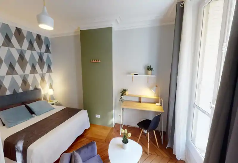 Camera da letto luminosa e stilosa con un ampio letto comodo, parete decorativa geometrica e una scrivania vicino alla finestra — ideale per riposo e lavoro leggero.