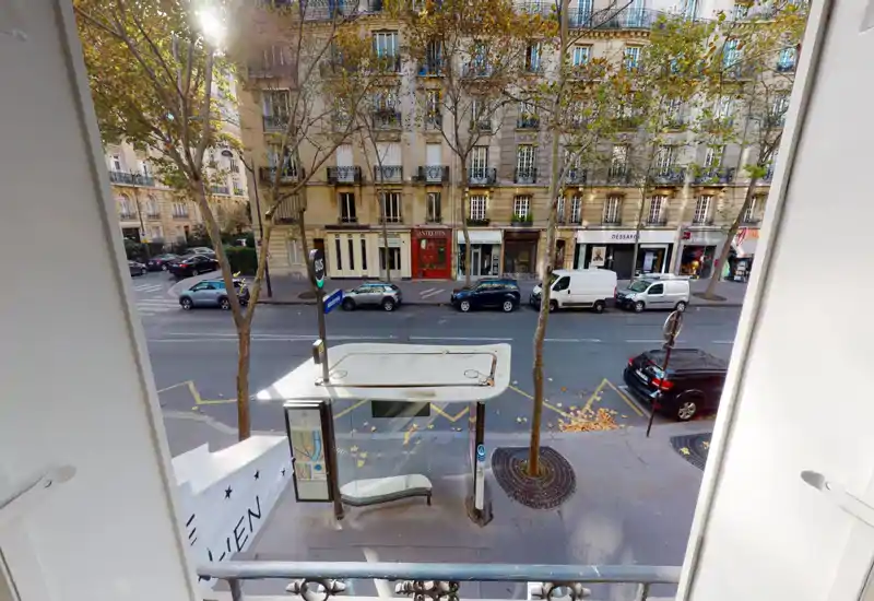 Vista sulla strada da una finestra o piccolo balcone con pensilina dell'autobus, auto parcheggiate e viale alberato in stile parigino—buona luce naturale e contesto urbano chiaro.