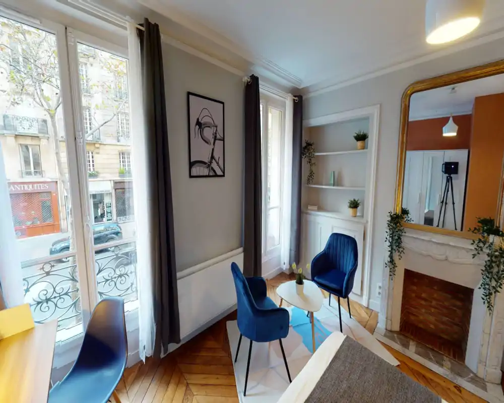Coin chambre lumineux et élégant avec grandes fenêtres, deux fauteuils en velours et une petite table basse près du lit ; parquet en chevrons et cheminée décorative.