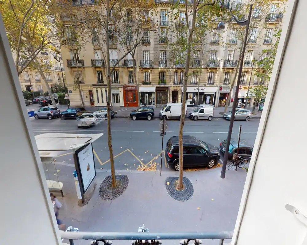 Vue sur la rue depuis une fenêtre/balcon au premier étage : avenue bordée d'arbres avec voitures garées, commerces et abribus — idéale pour présenter le quartier.
