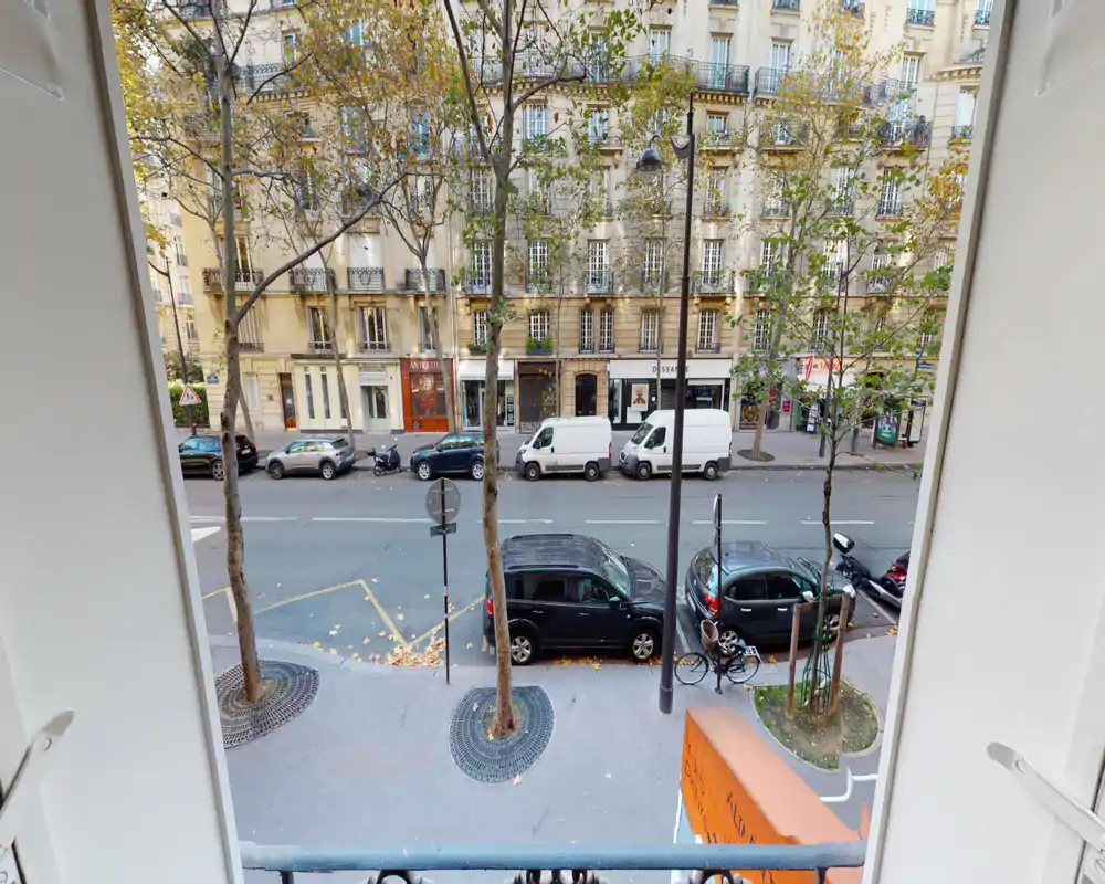 Street view from a window/balcony overlooking parked cars and classic Parisian façades; trees and sidewalk visible, offering a good urban outlook.