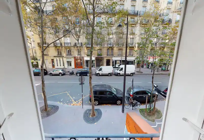 Street view from a window/balcony overlooking parked cars and classic Parisian façades; trees and sidewalk visible, offering a good urban outlook.
