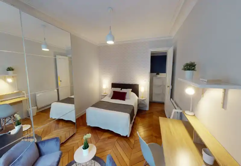 Bright, stylish bedroom with a double bed centered against a chevron-patterned accent wall, bedside lamps, mirrored wardrobe and a workspace. Warm wooden herringbone floor and thoughtful lighting create a welcoming, well-organized space.