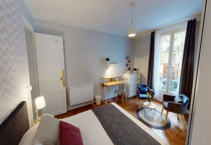 Comfortable, well-lit bedroom with a double bed, bedside lamp, desk and a cozy seating area by a tall window with curtains. Warm wooden flooring and tasteful decor create a welcoming atmosphere ideal for rental photos.