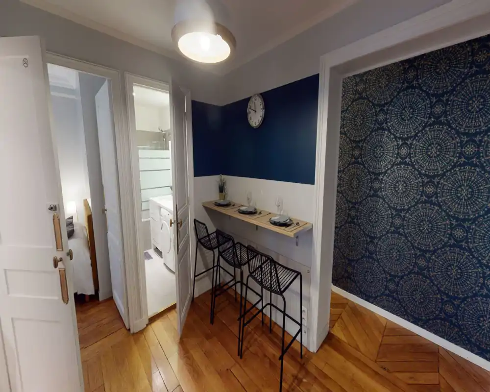 Compact living/dining corner with a wall-mounted bar table and three stylish stools, decorative patterned wallpaper and warm wooden floors — a bright, modern space ideal for casual meals.