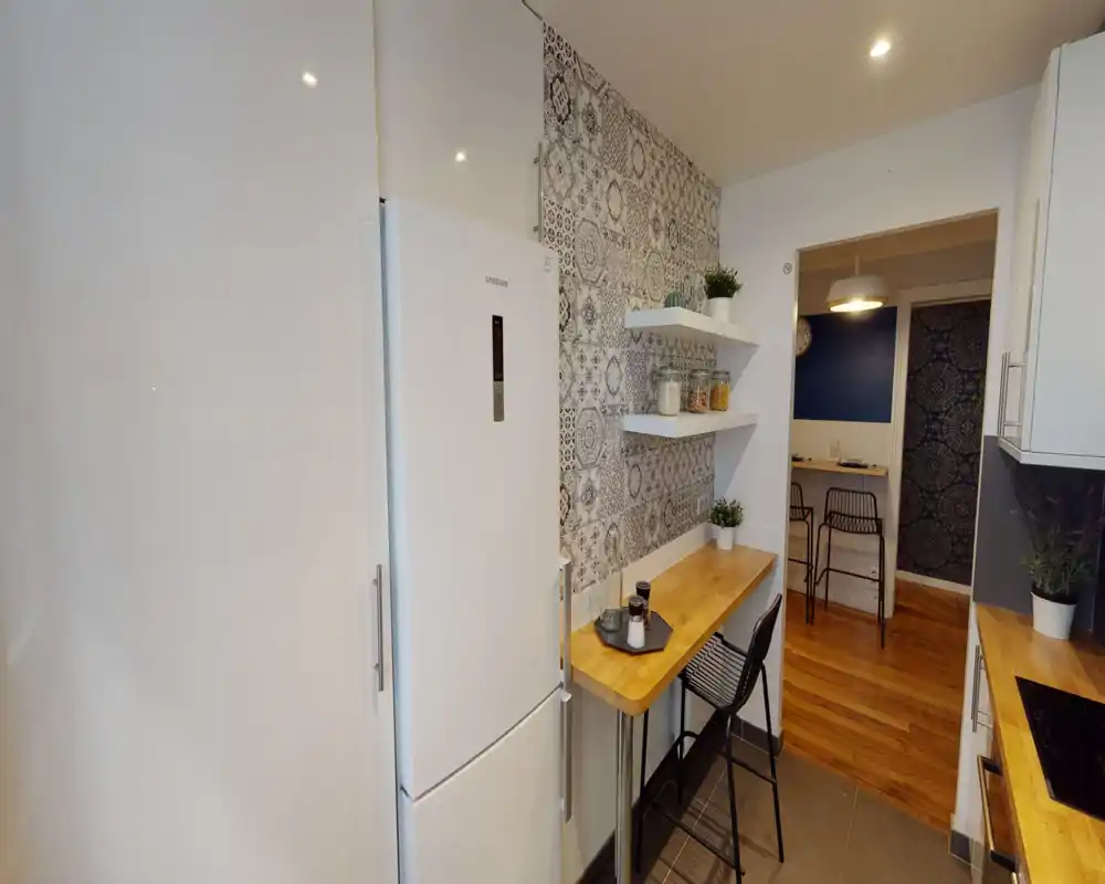 Compact, modern kitchen with wooden countertops, patterned feature wall and integrated fridge. A small breakfast bar with stools and open shelving creates a cozy, functional space.