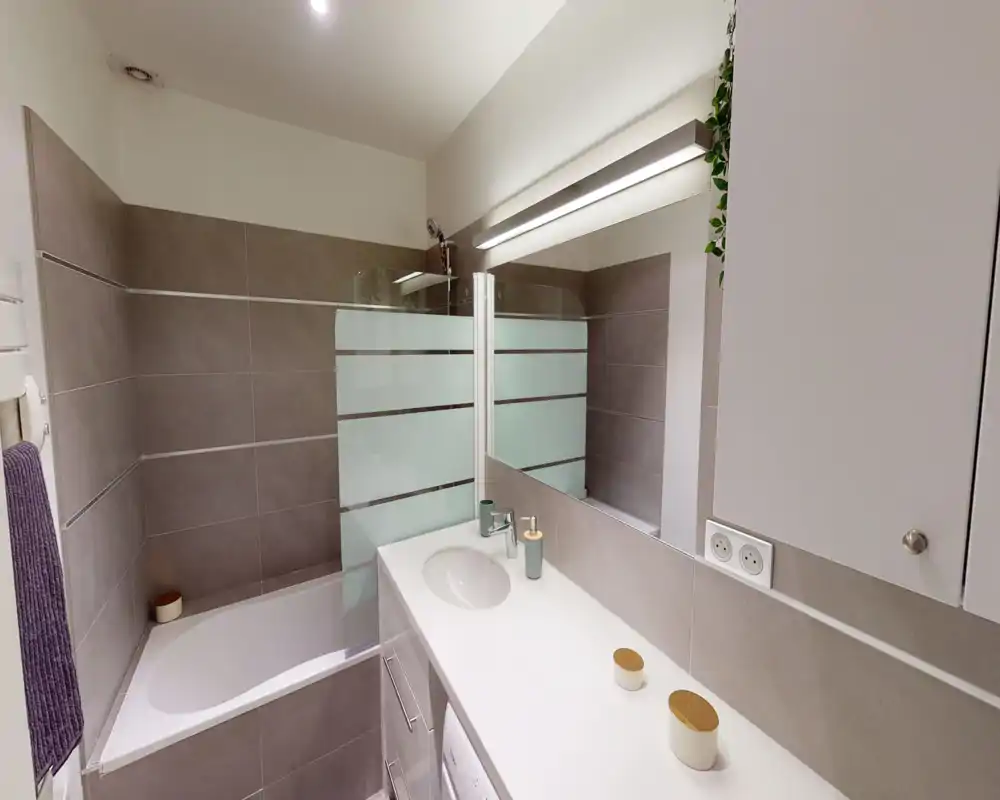 Modern tiled bathroom with a bathtub, partial glass shower screen and a wide vanity with sink and mirror. Neutral tones and clean surfaces create a fresh, well-lit space.