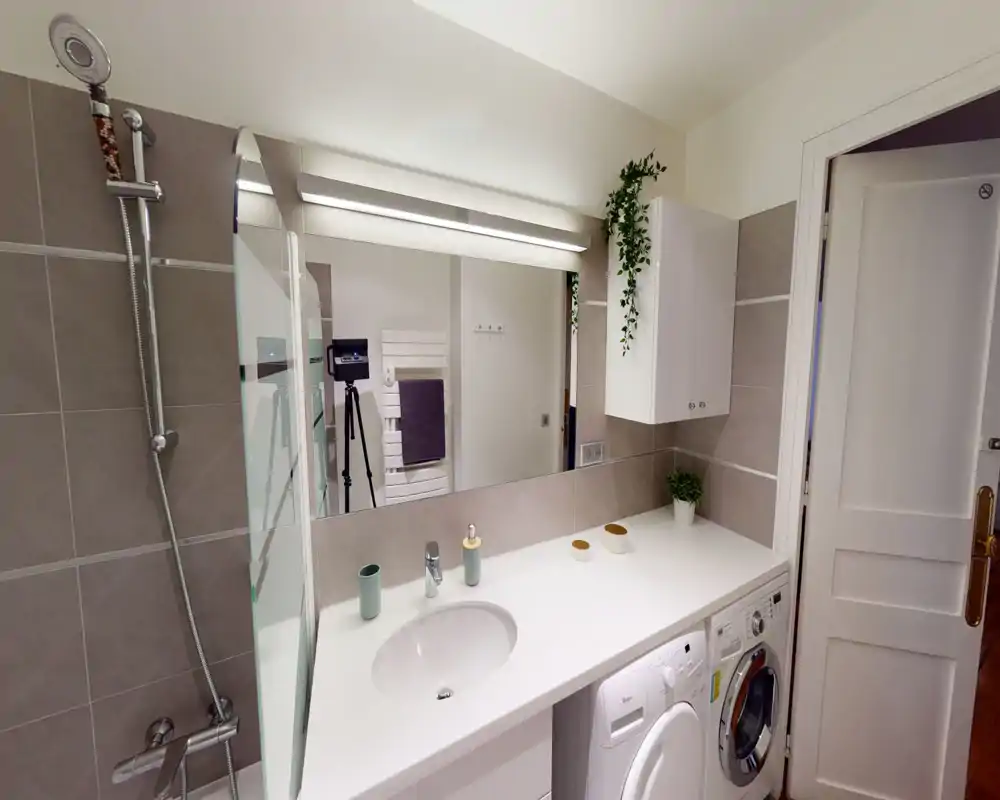 Bright, modern bathroom with a sink integrated into a long countertop, mirror with overhead lighting, shower fixtures and a stacked washer and dryer under the counter.