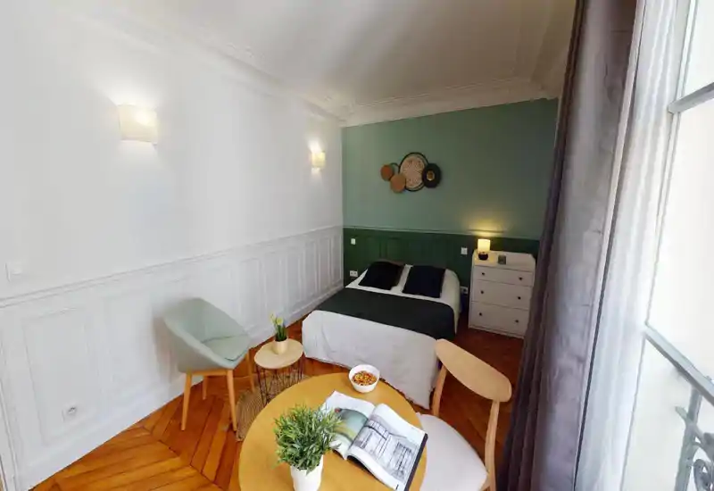Bright, styled bedroom with a double bed, bedside chest, and decorative wall paneling. Natural light from a large window and warm wooden floors create a cozy, modern feel — ideal for short-term rentals.