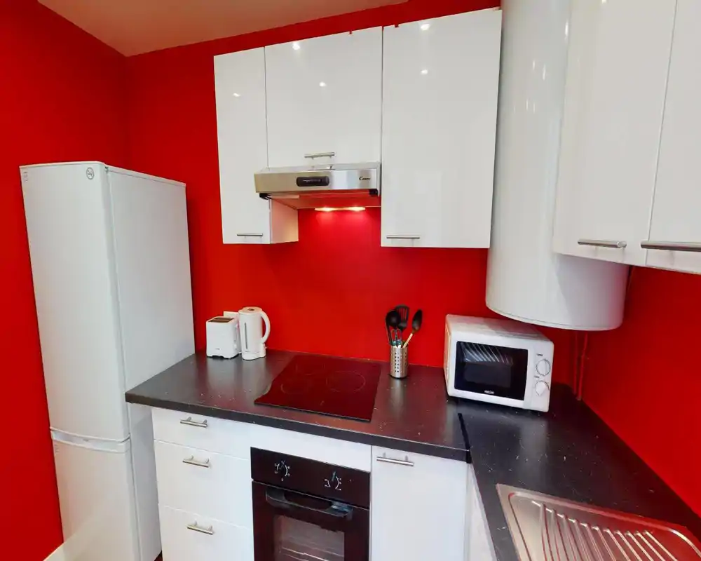Modern compact kitchen with white cabinets, electric hob and appliances set against a bold red wall — bright, tidy and well-equipped for short stays.