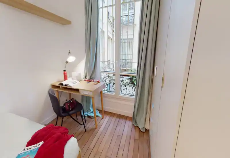 Small, bright bedroom with a single bed, wooden floors, a study desk by a tall window and built-in wardrobe; tasteful, airy styling ideal for a student or single tenant.
