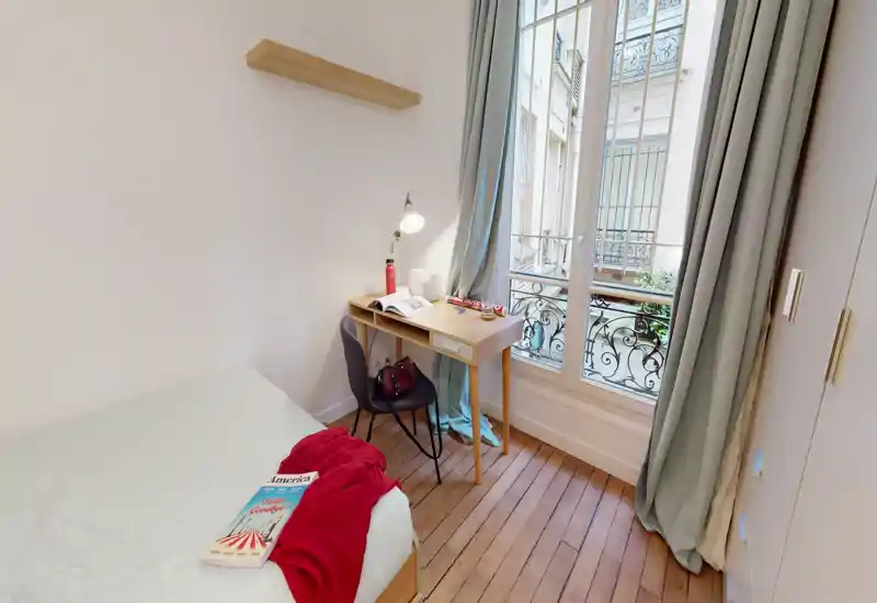 Bright, compact bedroom with a single bed, wooden floors and a small work desk by a tall window letting in abundant natural light.