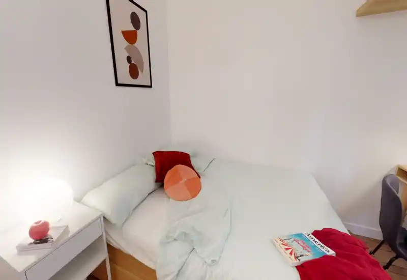 Cozy, well-lit bedroom with a double bed dressed in pale bedding, decorative cushions and a bedside lamp; simple modern decor and a reading book on the bed.