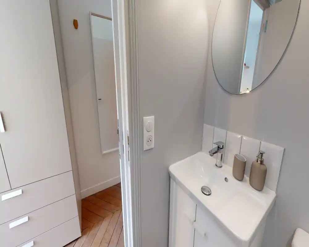 Compact, modern bathroom corner with a white sink, chrome faucet, round mirror and neutral-tone soap dispensers — clean and bright.