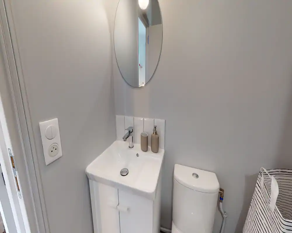 Compact, modern bathroom corner with a white sink, faucet, round mirror and a close-coupled toilet; neutral grey walls and minimal accessories create a clean, functional look.