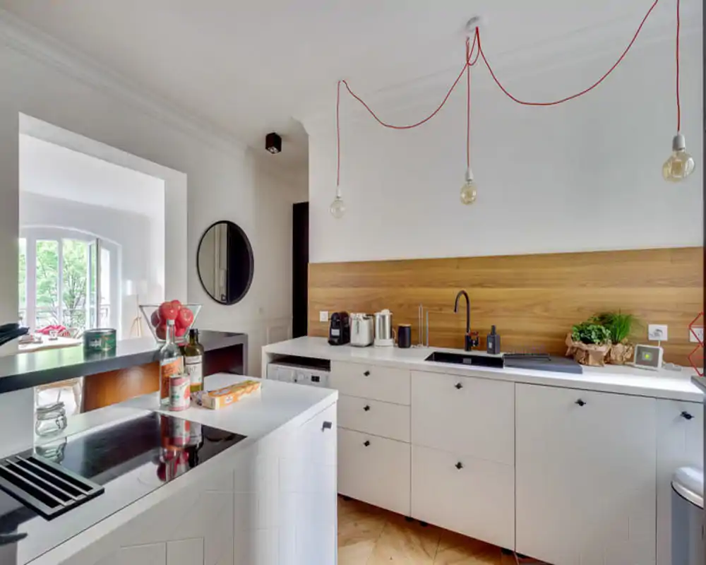 Modern, bright kitchen with white cabinets, wooden backsplash and integrated island. Stylish pendant bulbs and natural light create a warm, welcoming cooking area.