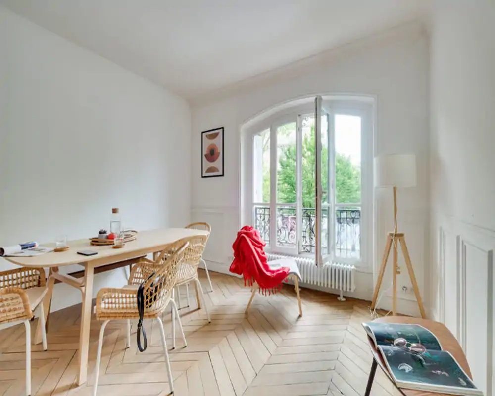 Bright, elegant dining area with a wooden table, woven chairs and a large arched window letting in abundant natural light. The red throw on a lounge chair and tasteful minimal decor create a warm, welcoming atmosphere.