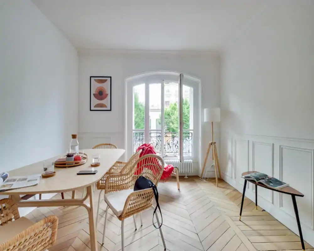 Bright, stylish dining area with a light-wood table, wicker chairs and a large arched window letting in abundant natural light.