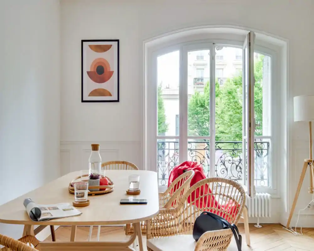 Sunny, styled dining area with a wooden table, woven chairs and a large arched window opening to a small balcony — bright and inviting for meals.