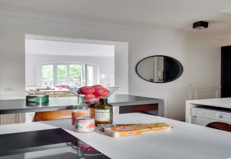 Bright modern kitchen corner with a countertop island showing pantry items and a bowl of apples; natural light from a large arched window and clean, minimal white walls.