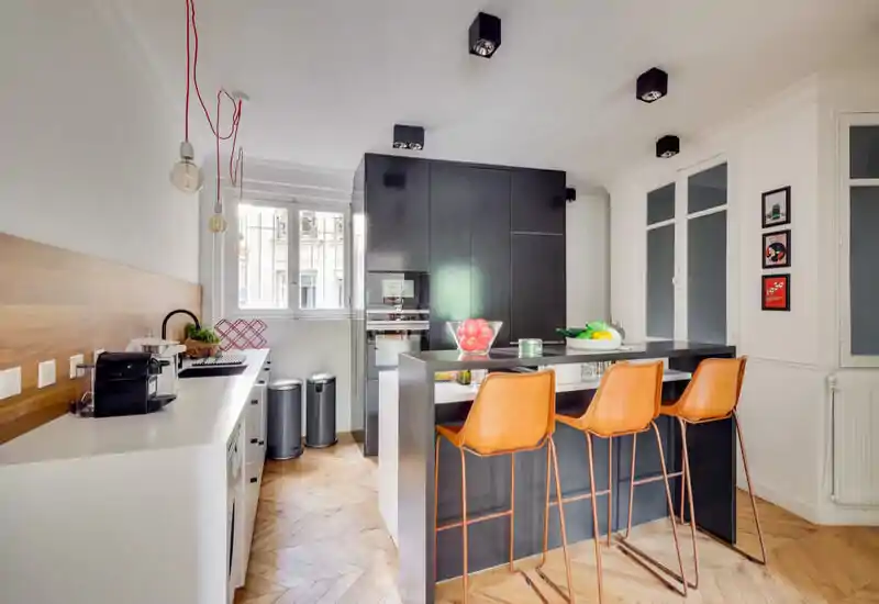 Bright modern kitchen with a central island and three leather bar stools, black cabinetry and warm wooden herringbone floor — ideal for social cooking and casual dining.