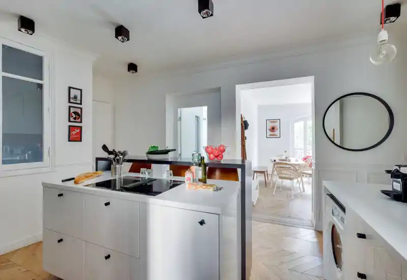 Bright modern kitchen with a central island, built-in cooktop and white cabinetry, flowing into a dining area; warm wood herringbone floors and tasteful styling create a welcoming, functional space.