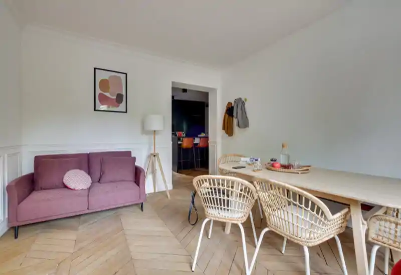 Bright, stylish living/dining area with a pink two-seater sofa, rattan dining chairs and a large wooden table — calm neutral walls and parquet herringbone flooring add warmth and charm.