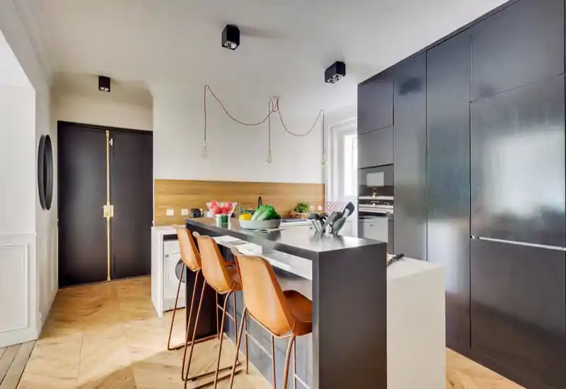 Modern open kitchen with a central island, three leather bar stools and integrated tall cabinetry; warm wood flooring and natural light create a stylish, functional cooking and dining area.