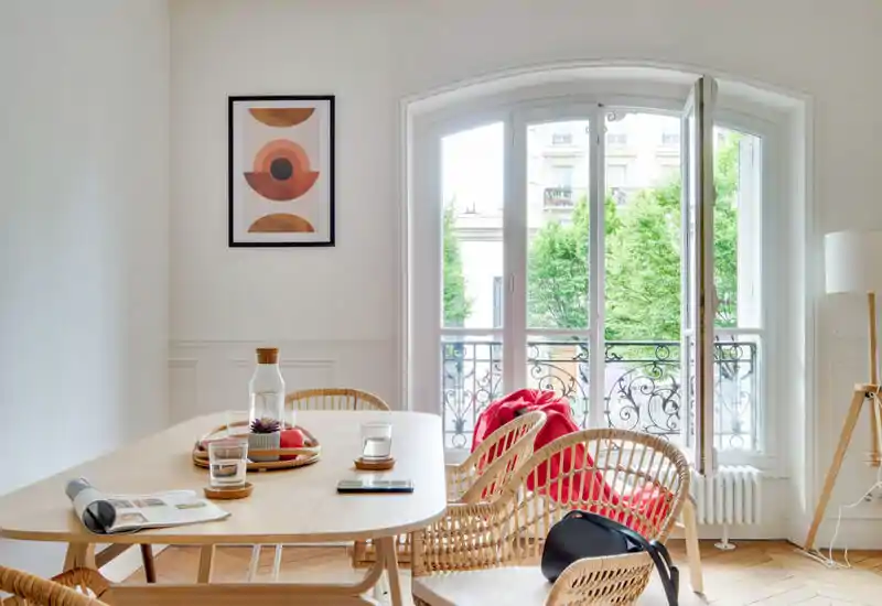 Sunny, styled dining area with a wooden table, woven chairs and a large arched window opening to a small balcony — bright and inviting for meals.