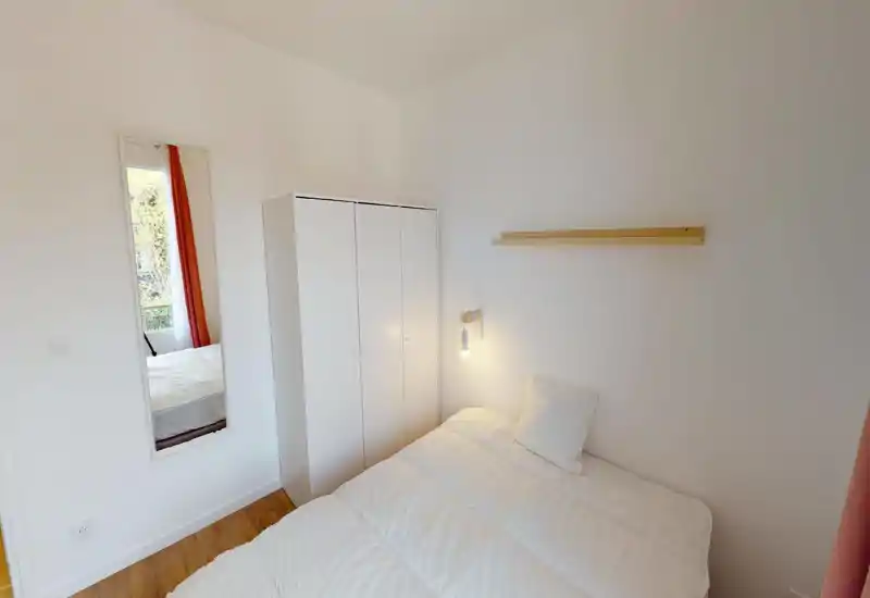 Camera da letto luminosa e minimal con un comodo letto matrimoniale, armadio bianco e una lampada da lettura a parete. Pavimento in legno, specchio e molta luce naturale.