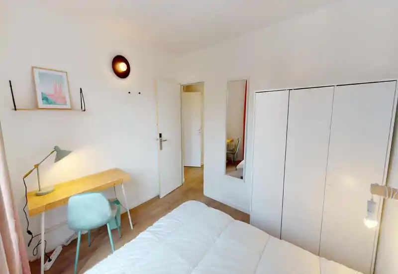 Camera da letto luminosa e ben arredata con letto comodo in primo piano, scrivania in legno e sedia pastello per lavorare e un grande armadio bianco per riporre gli abiti.