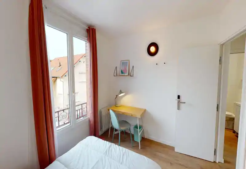 Camera da letto luminosa e ordinata con un letto ben visibile in primo piano, una scrivania in legno e una sedia vicino alla finestra e tende color corallo che lasciano entrare molta luce.