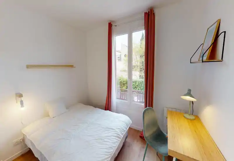 Camera da letto luminosa e compatta con letto matrimoniale, scrivania in legno e finestra con tende rosse che lasciano entrare luce naturale.