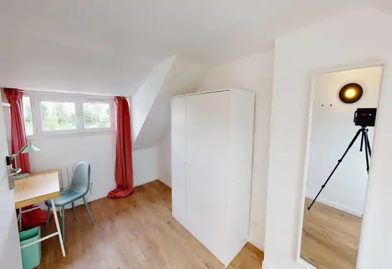 Piccola camera mansardata/ufficio con soffitto inclinato, ampia armadio bianco, scrivania in legno e sedia vicino a una finestra ampia con tende rosse che lasciano entrare luce naturale.