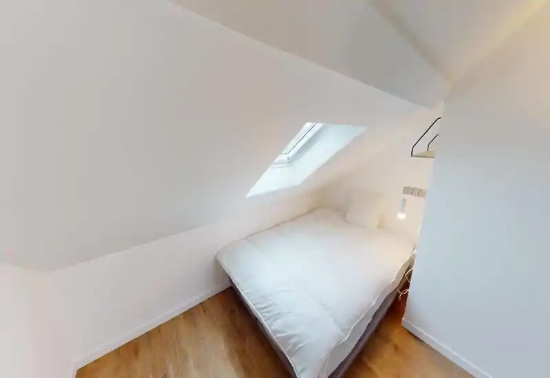 Camera da letto mansardata compatta con un letto matrimoniale vicino a una finestra a tetto, luce naturale intensa e arredi bianchi e minimalisti.