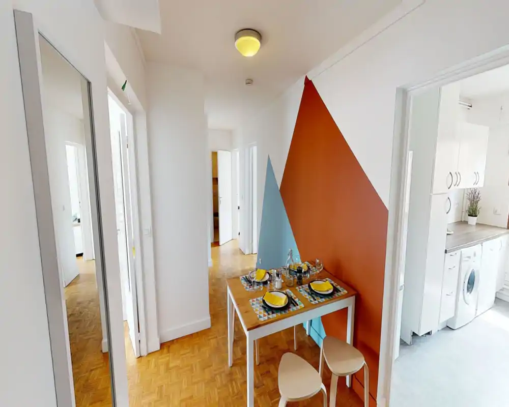 Bright hallway/entrance with parquet flooring, a small styled dining table against a colorful geometric feature wall and a tall mirror; the kitchen is visible through an open doorway.