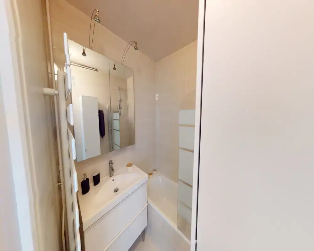 Compact modern bathroom with a white vanity, sink, mirror cabinet and a bathtub with partial glass screen. Neutral beige tiles and simple fixtures create a clean, functional space.